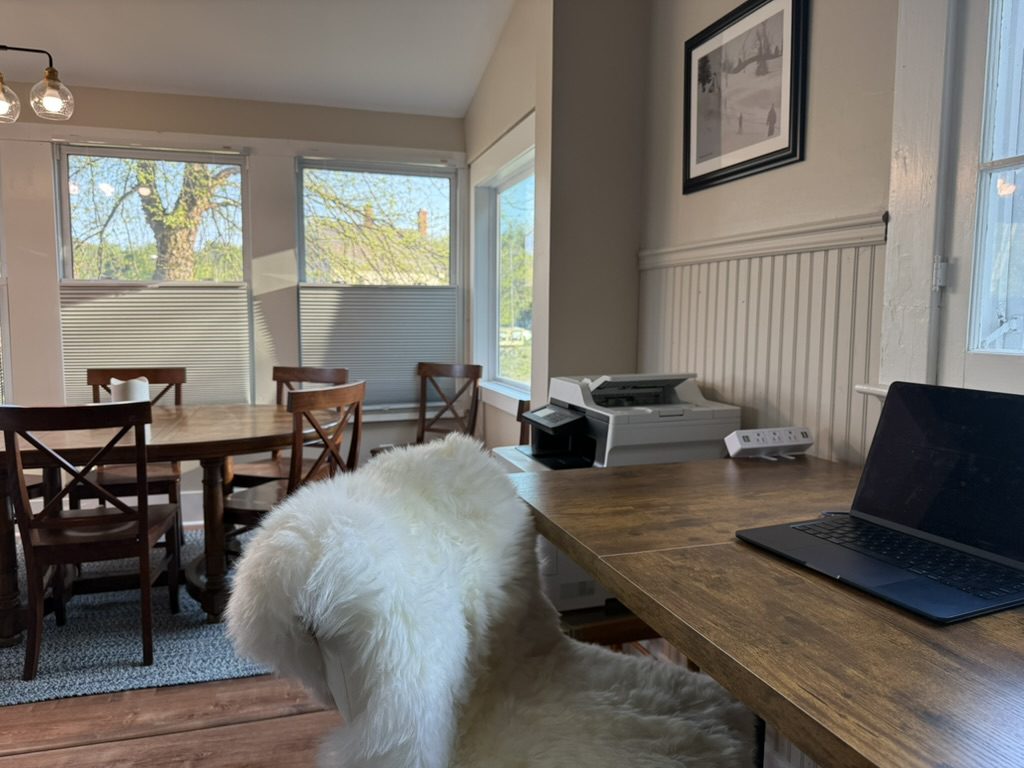 The standing desk at Eiswork under the windows, with a laptop, sheepskin-draped chair and the printer behind