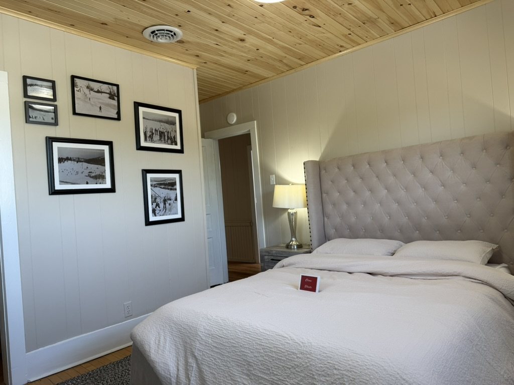 Bedroom in the Eiswork Loft at 235 Main Street — knotty pine ceiling, white panelled walls, a king bed and a wall of framed vintage North Creek ski photographs