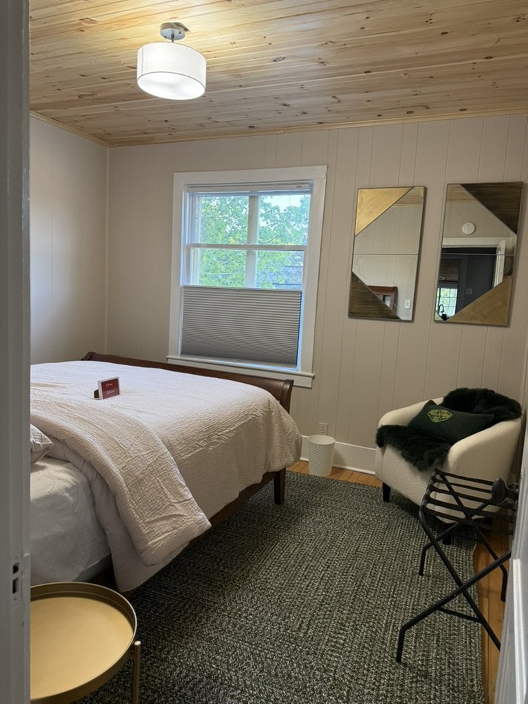 Second bedroom in the Eiswork Loft at 235 Main Street — pine ceiling, sleigh bed, green wool rug and a bouclé armchair by the window