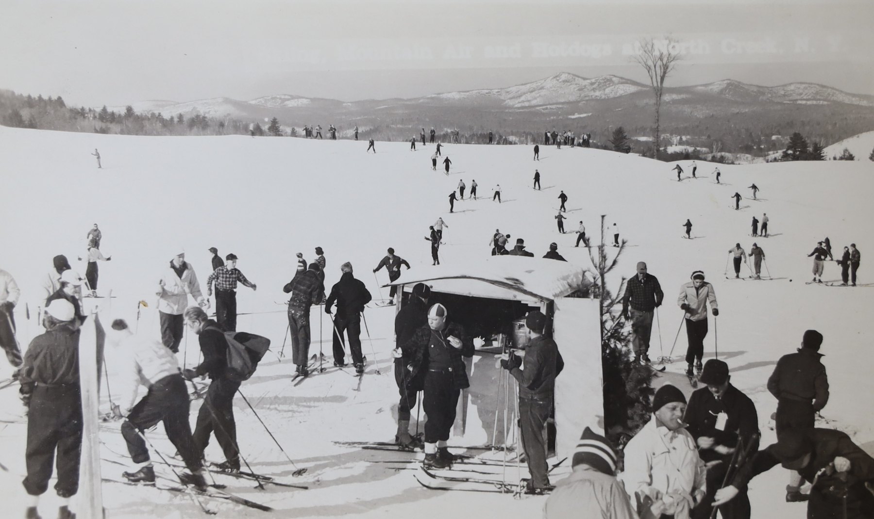 Skiers gathered around a hot dog stand on the slope at the North Creek Ski Bowl in the 1930s, with the Adirondack range behind them